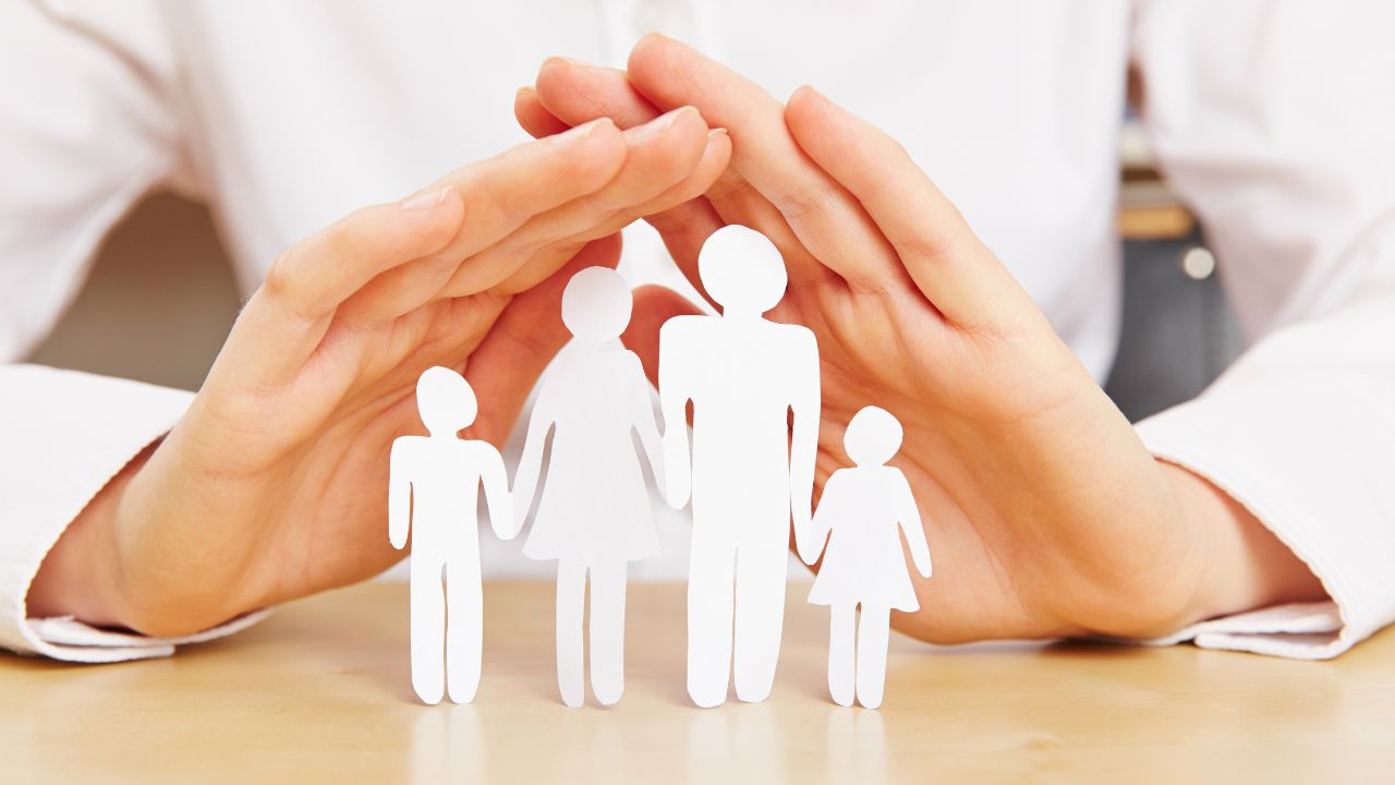 Über einer aus Papier ausgeschnittenen Familie aus Eltern und zwei Kindern werden zwei schützende Hände gehalten.