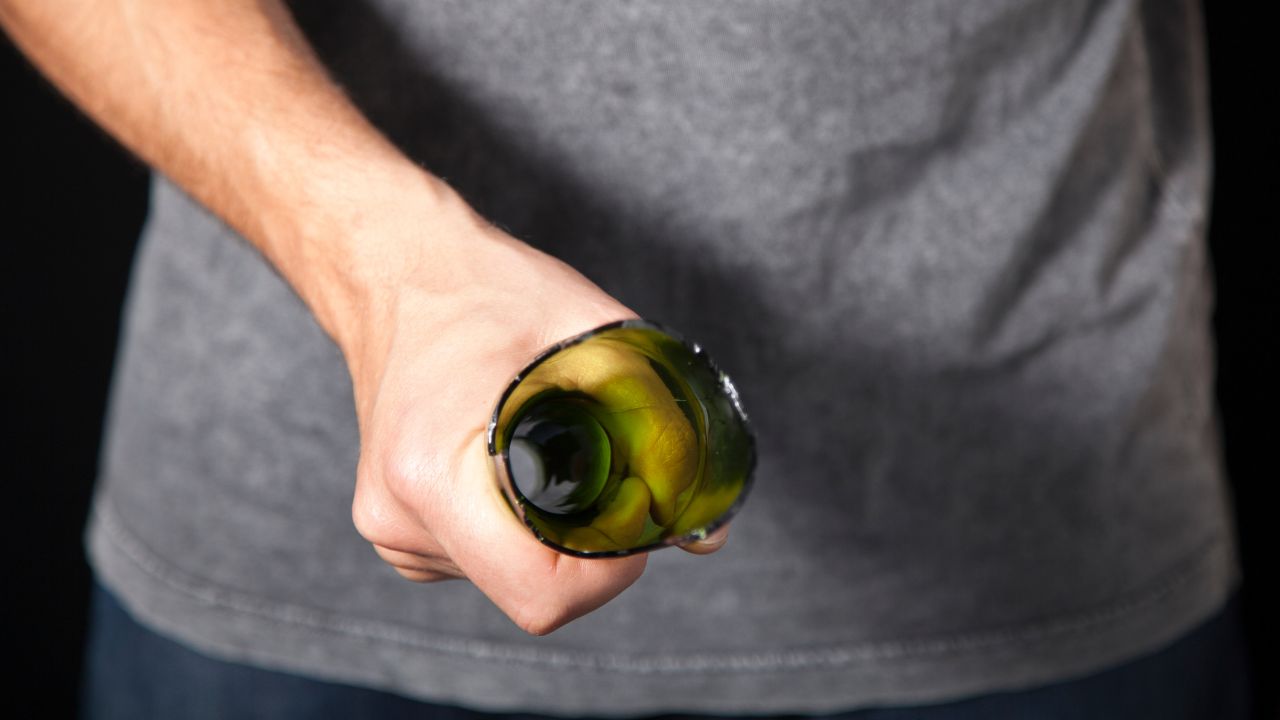 Mann in grauem T-Shirt hält eine abgebrochene grüne Glasflasche mit scharfen Kanten in der Hand.	