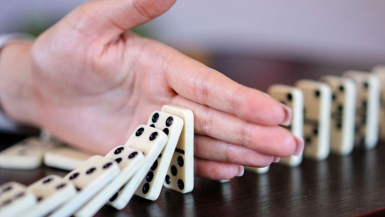 Nahaufnahme einer Hand, die fallende Dominosteine aufhält, während die restlichen Steine noch stehen.