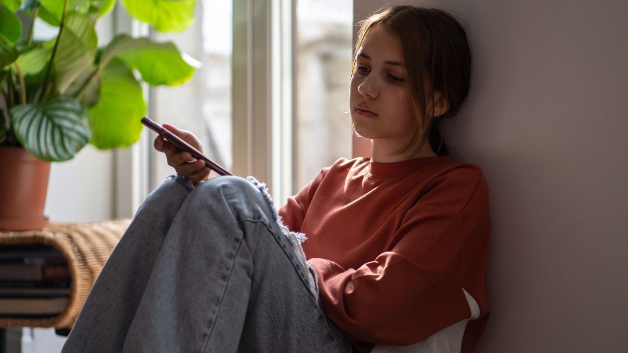 Mädchen in rostrotem Pullover und Jeans sitzt an einer Wand neben einem Fenster und schaut auf ihr Smartphone.