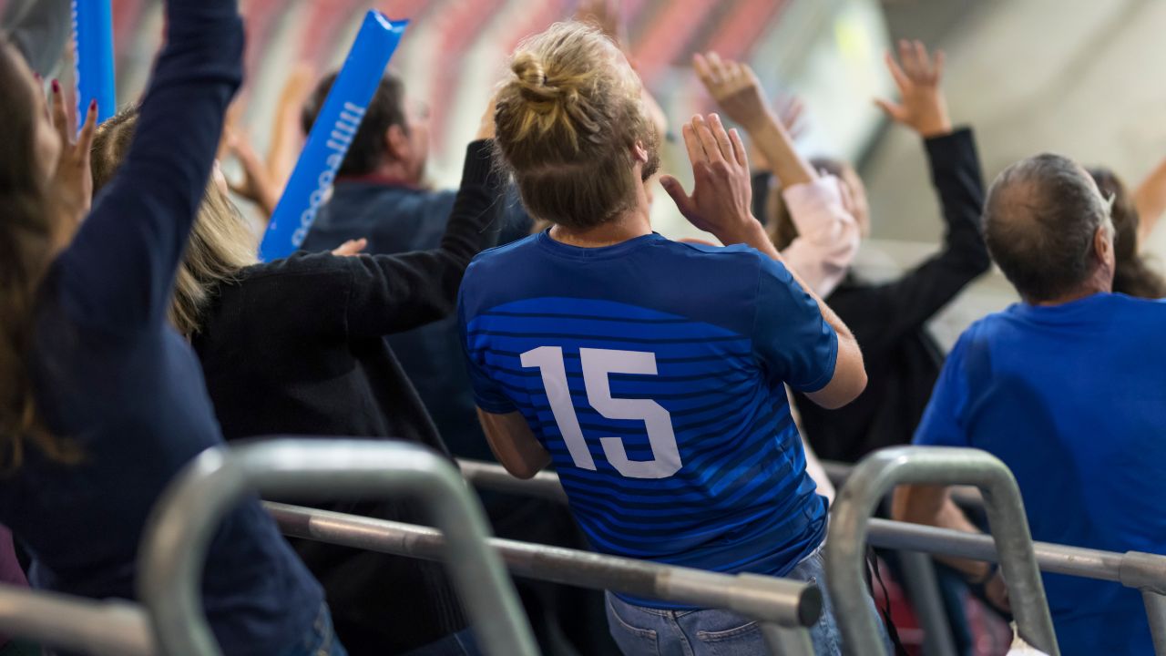 Jubelnde Fans in einem Stadion, im Fokus ein Mann in einem blauen Trikot mit der Nummer 15 von hinten.