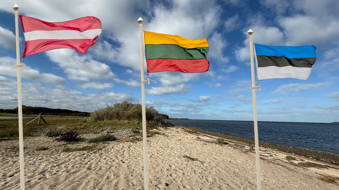 Die Flaggen der baltischen Staaten – Lettland, Litauen und Estland – wehen an weißen Fahnenmasten auf einem Sandstrand neben dem Meer unter einem blau-weißen Wolkenhimmel.