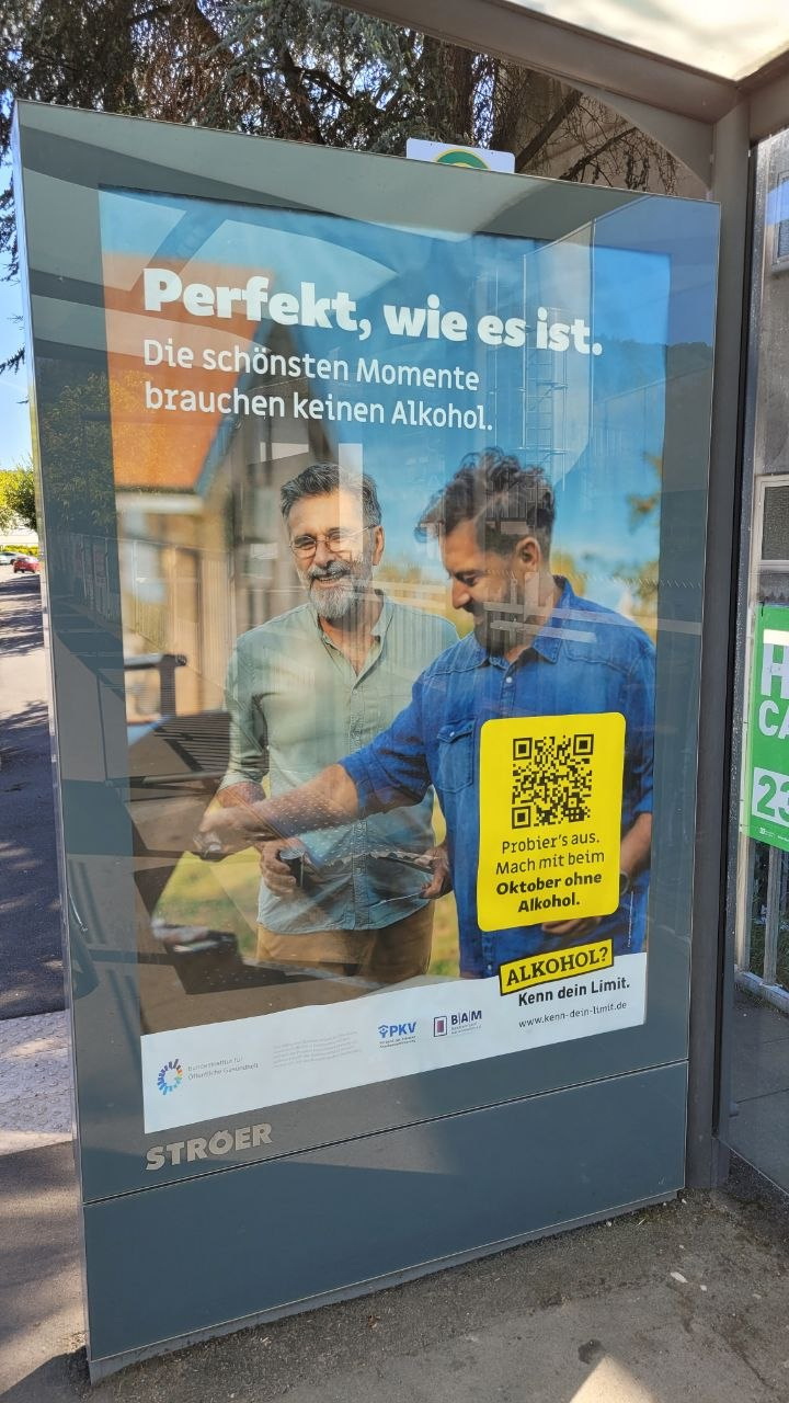 Werbeplakat an einer Bushaltestelle für die Kampagne 'Kenn dein Limit': Zwei lächelnde Männer stehen draußen zusammen. Große Überschrift: 'Perfekt, wie es ist. Die schönsten Momente brauchen keinen Alkohol.' Daneben ein gelber Kasten mit QR-Code und dem Text 'Probier’s aus. Mach mit beim Oktober ohne Alkohol.' Unten steht 'ALKOHOL? Kenn dein Limit.' mit dem dazugehörigen Logo und der Webadresse.