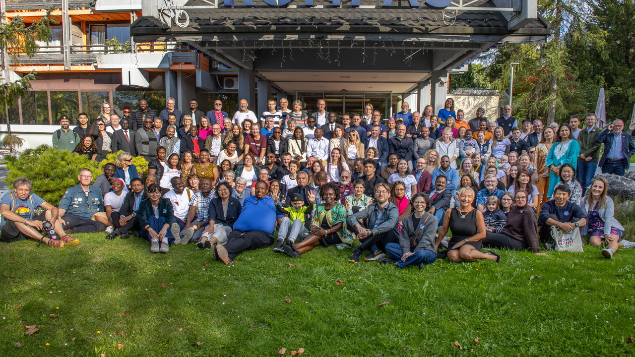  Gruppenfoto von über hundert Menschen unterschiedlichen Alters und Hintergrunds, die sich im Freien vor einem Hotel mit überdachtem Eingang versammeln. Vorne sitzen einige auf dem Rasen.