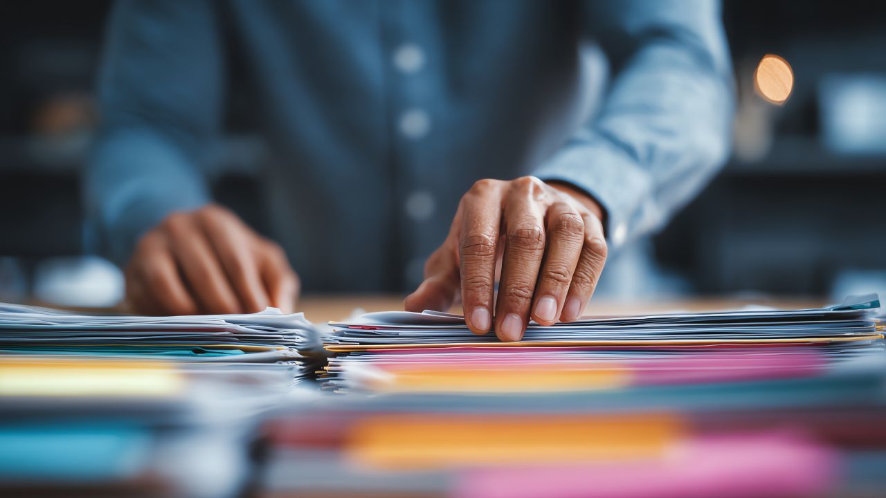 Nahaufnahme von zwei Händen einer Person im hellblauen Hemd, die auf einem Schreibtisch liegende Stapel bunter Dokumente und Ordner sortiert.