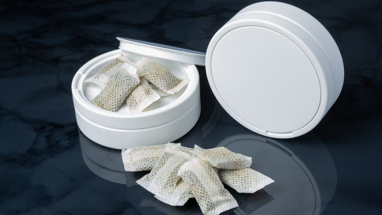 SGeöffnete weiße Runddose mit Nicotinbeuteln (Snus-Pouches) darin und einige Beutel davor auf dunkler Marmoroberfläche. Der Deckel steht angelehnt dahinter.