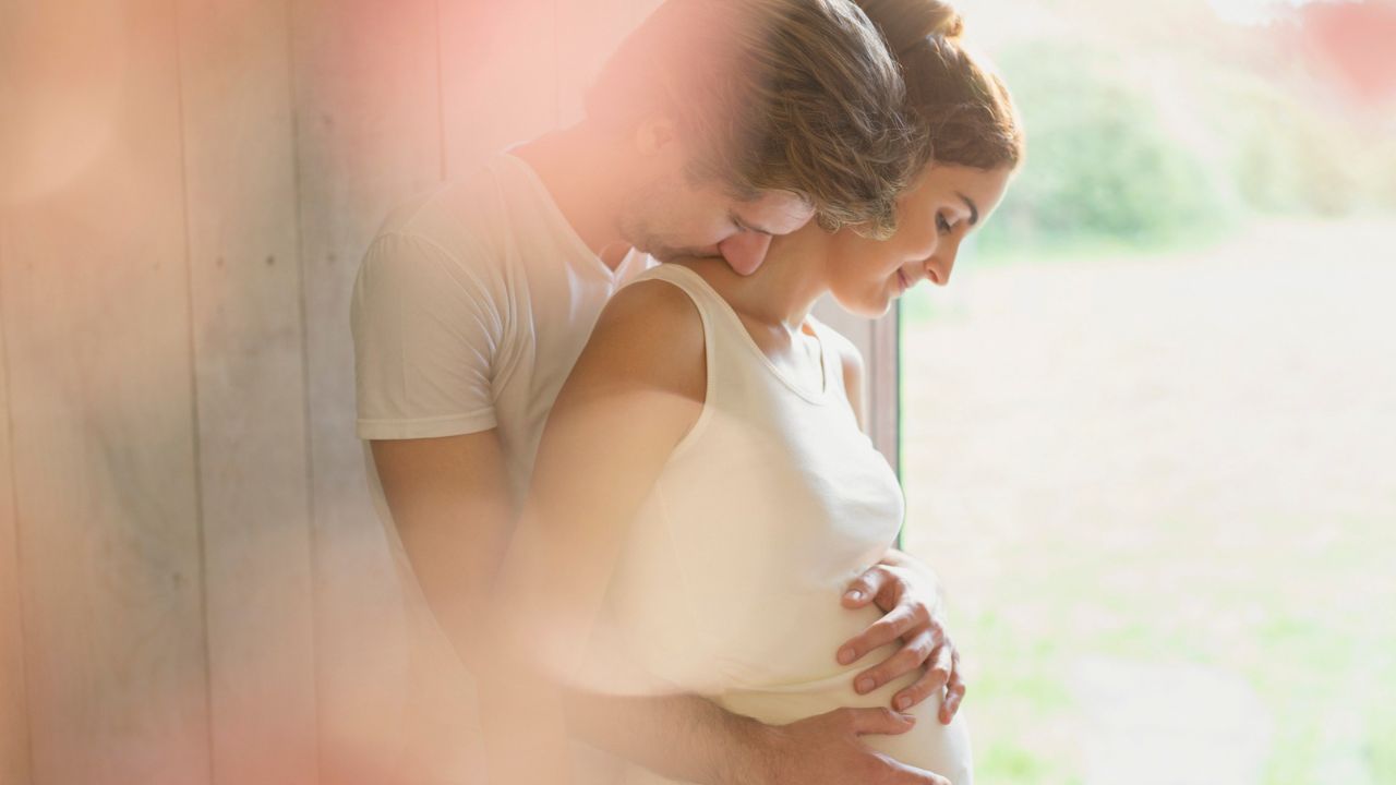 Schwangere Frau in weißem Top wird von ihrem Partner von hinten umarmt. Beide halten liebevoll ihren Babybauch, während er sie am Hals küsst. Inniger Moment in hellem Sonnenlicht vor einer Holzwand.