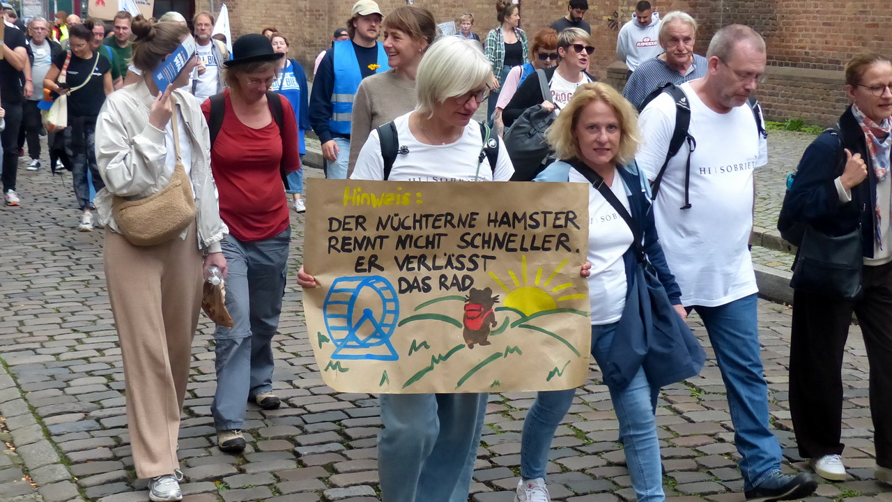 Gruppe von Menschen bei einer Demonstration auf einem gepflasterten Weg. Im Vordergrund hält eine Frau ein handgemaltes Schild mit einer humorvollen Botschaft in deutscher Sprache: 'Hinweis: Der nüchterne Hamster rennt nicht schneller. Er verlässt das Rad.' Das Schild zeigt eine Zeichnung eines Hamsterrads mit einem Hamster, der es verlässt, und einer Sonne im Hintergrund. Die Teilnehmer*innen tragen Alltagskleidung und wirken entspannt.