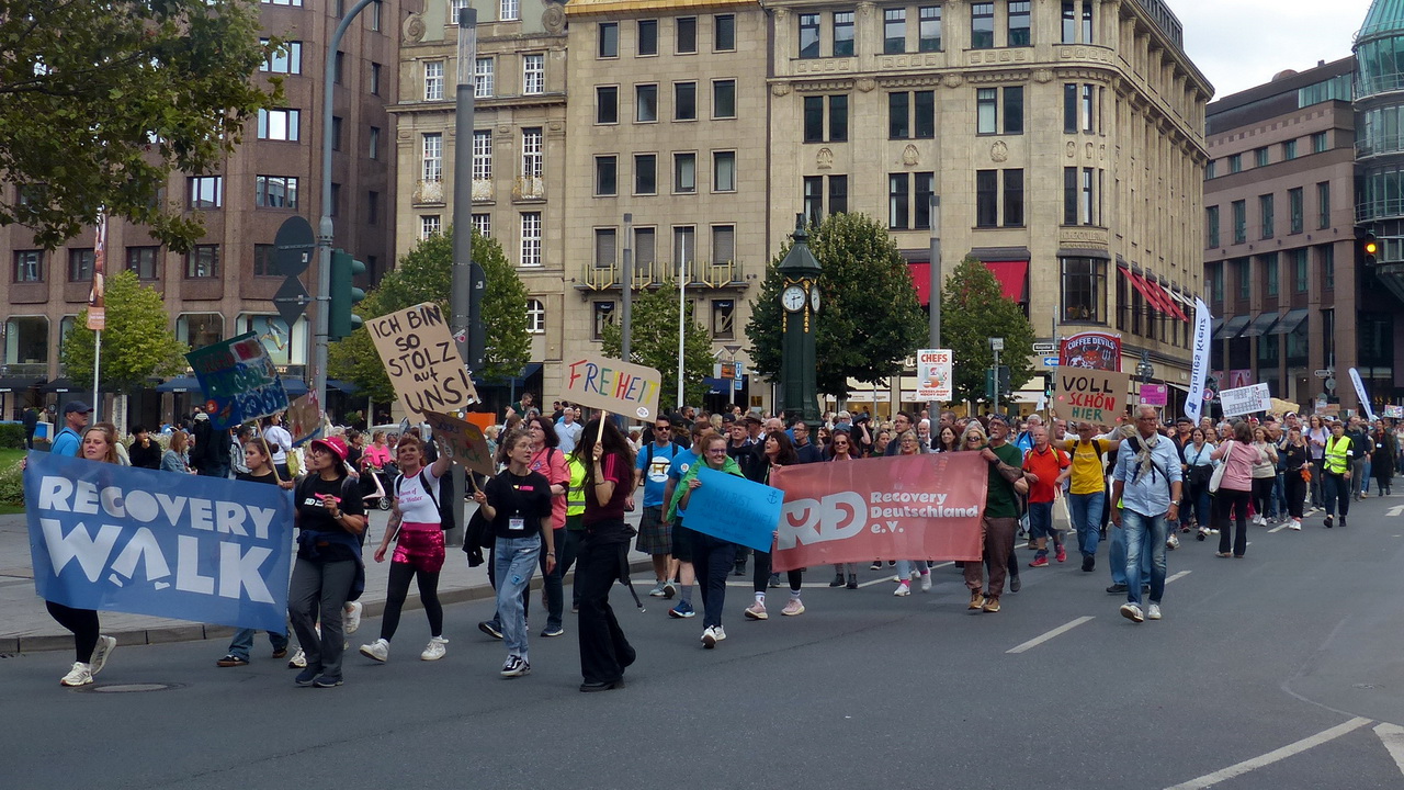 Ein Gruppe von Demonstrant*innen zieht bei einer 'Recovery Walk'-Veranstaltung durch eine Innenstadtstraße. Sie tragen transparente und handgeschriebene Schilder mit Botschaften wie 'Ich bin so stolz auf uns', 'Freiheit' und 'Voll schön hier', sowie ein großes blaues Banner mit der Aufschrift 'Recovery Walk'. Im Vordergrund wird auch ein rotes Banner des 'Recovery Deutschland e.V.' gezeigt. Die Menge ist bunt gemischt und schreitet an alten, mehrstöckigen Gebäuden vorbei.