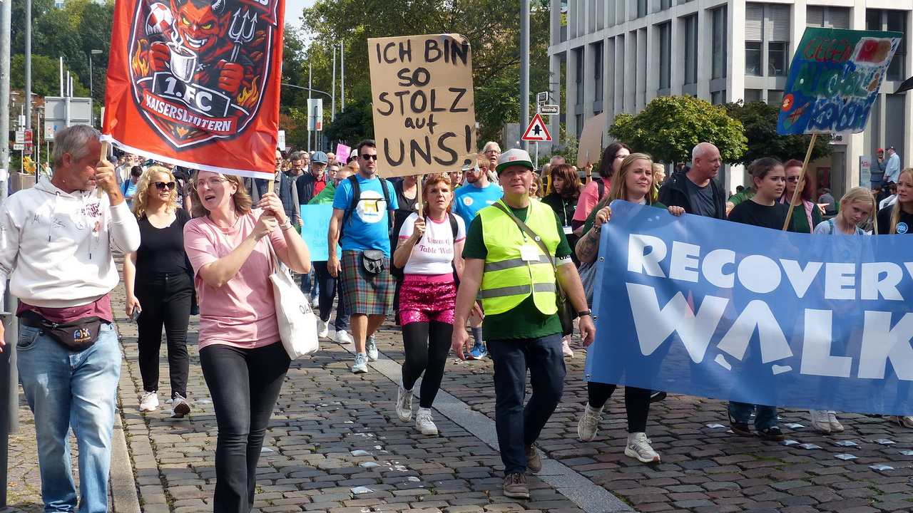 Teilnehmer*innen eines 'Recovery Walk' marschieren über eine gepflasterte Straße. Sie tragen ein großes blaues Banner mit der Aufschrift 'RECOVERY WALK', eine rote Flagge des 1. FC Kaiserslautern und ein handgeschriebenes Pappschild mit dem Text 'ICH BIN SO STOLZ auf UNS!'.