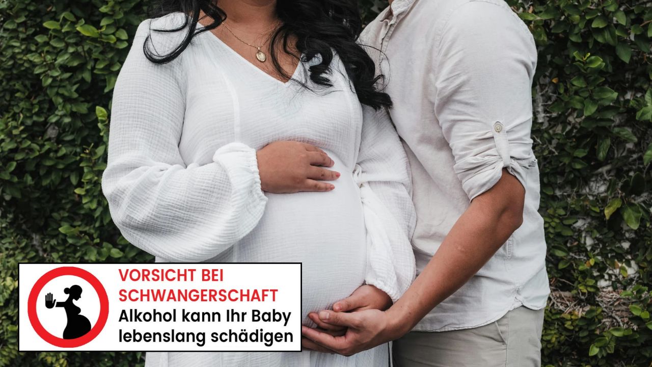 Eine schwangere Frau in einem weißen Kleid und ihr Partner halten gemeinsam ihren Babybauch vor einer grünen Pflanzenwand. Unten links ist ein Warnhinweis eingeblendet: 'VORSICHT BEI SCHWANGERSCHAFT – Alkohol kann Ihr Baby lebenslang schädigen'.