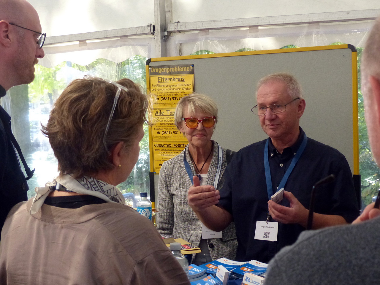 Gruppe von Personen an einem Informationsstand unter einem Festzelt. Im Mittelpunkt stehen eine Frau mit orangefarben getönter Brille und ein älterer Mann mit Lanyard, die im Gespräch mit Besucher*innen sind. Auf dem Tisch liegen Informationsbroschüren.