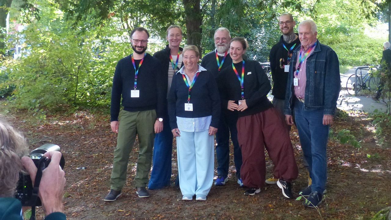 Gruppenfoto von sieben Personen unterschiedlichen Alters, die nebeneinander unter Bäumen auf laubbedecktem Boden stehen und lächelnd in die Kamera blicken. Alle tragen regenbogenfarbene Schlüsselbänder mit Namensschildern. Im Vordergrund links ist eine Person von hinten zu sehen, die die Gruppe fotografiert. Im Hintergrund grüne Sträucher, Bäume und abgestellte Fahrräder.