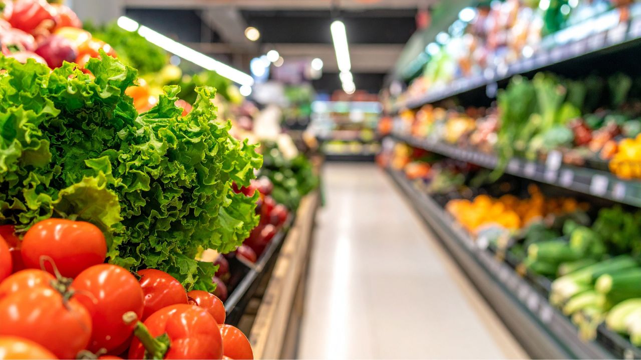 Frische Gemüseabteilung im Supermarkt mit grünem Blattsalat und roten Tomaten im Vordergrund, im Hintergrund unscharf weitere Obst- und Gemüseregale mit bunter Auswahl.
