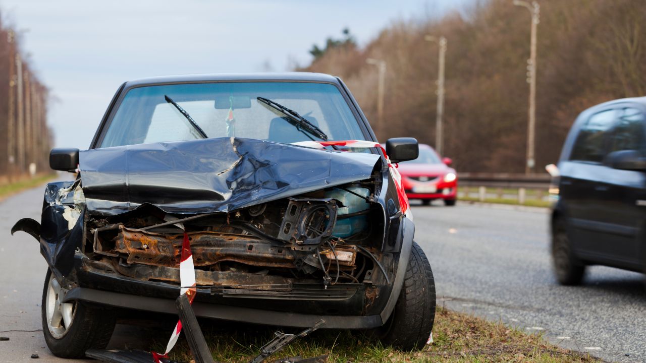 Ein stark beschädigtes dunkles Auto mit zerstörter Front steht am Straßenrand und ist mit rot-weißem Absperrband markiert. Im Hintergrund sind weitere Fahrzeuge auf einer Straße zu sehen.