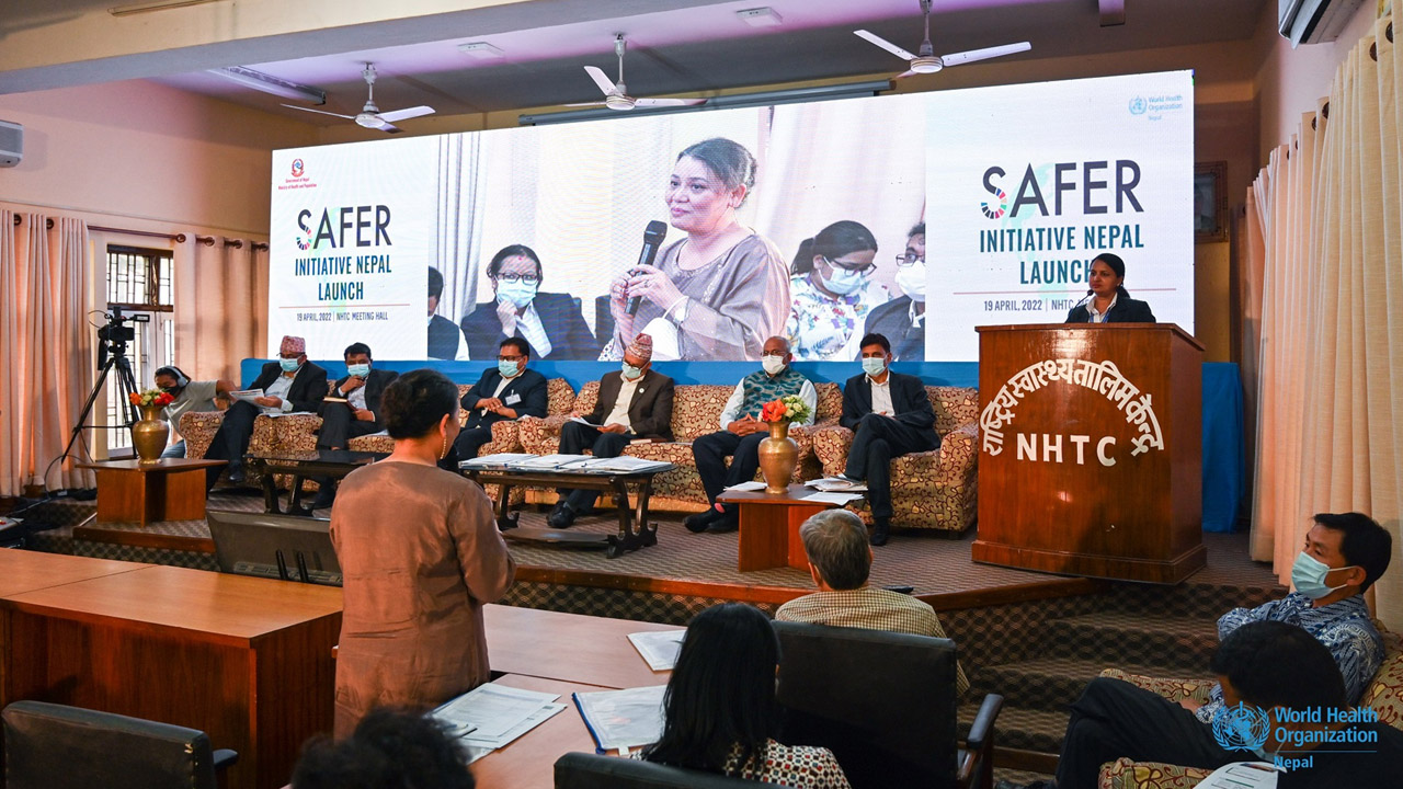 Eine Frau spricht mit einem Mikrofon während der Eröffnungsveranstaltung der 'SAFER-Initiative Nepal' am 19. April 2022 im NHTC-Sitzungssaal. Auf einem Podium sitzen mehrere Männer in Anzügen, während im Hintergrund eine Großleinwand die Rednerin zeigt und das Veranstaltungslogo präsentiert. Im Vordergrund verfolgt das Publikum die Präsentation.