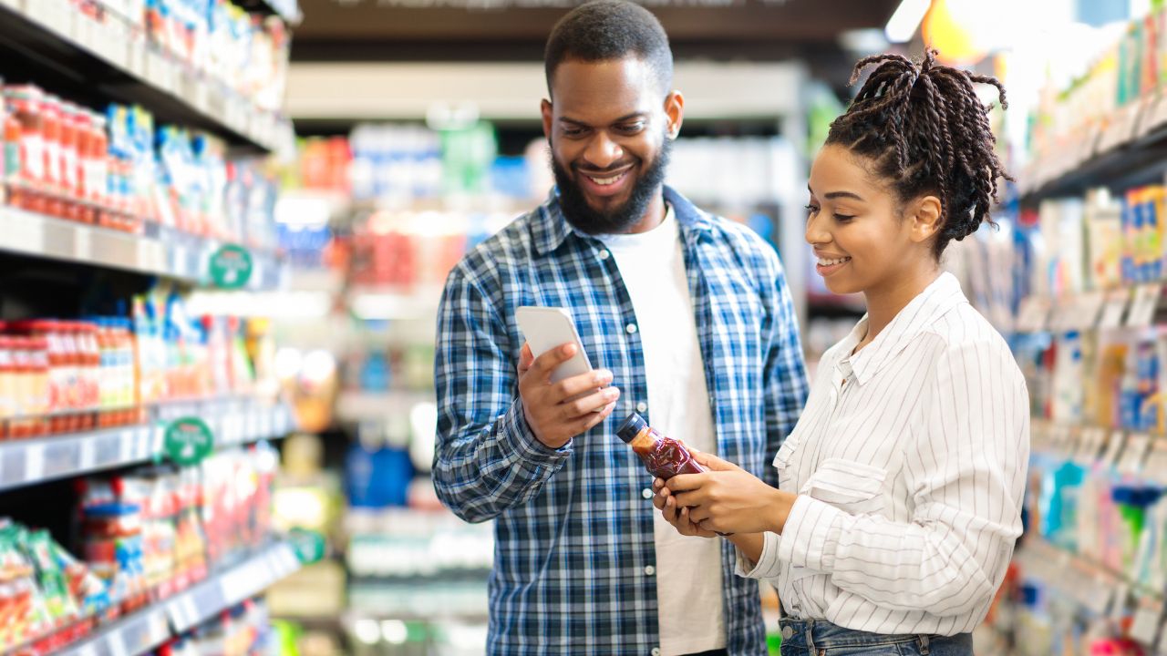 Mann und Frau beim Einkauf in einem Gang zwischen zwei Supermarktregalen betrachten ein Produkt und ein Smartphone in ihren Händen.