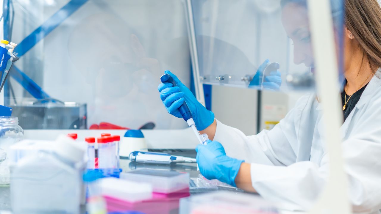 Eine Wissenschaftlerin in einem weißen Laborkittel und blauen Einweghandschuhen arbeitet in einem Labor. Sie hält eine Pipette und gibt eine Flüssigkeit in ein Reagenzglas. Im Vordergrund sind verschiedene Laborutensilien zu sehen, darunter Reagenzgläser mit roten Verschlüssen und eine Pipettenstation. Die Szene wird durch eine Schutzscheibe getrennt.