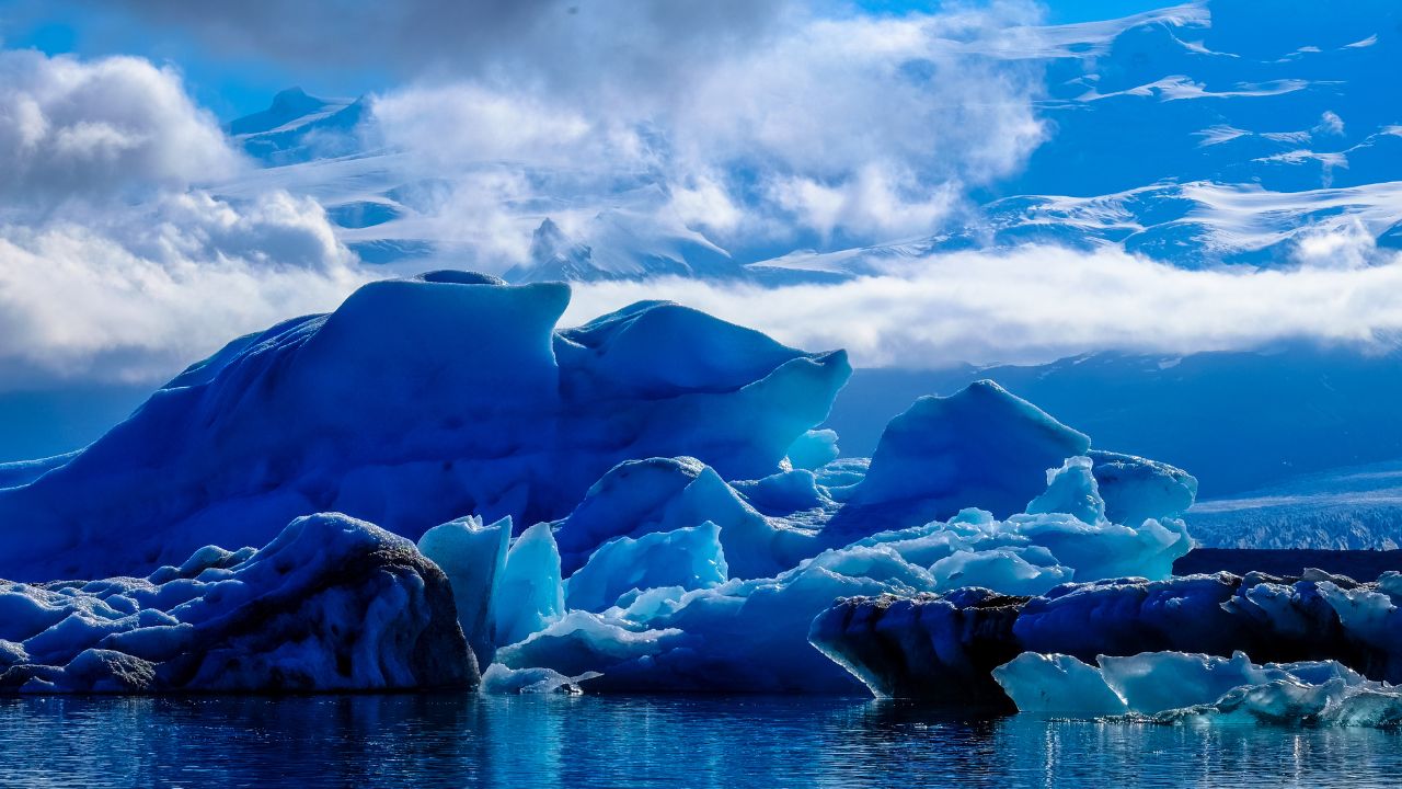 Eine majestätische Eislandschaft mit großen Eisbergen und Gletschern, die in einem tiefblauen Gewässer schwimmen. Im Hintergrund sind schneebedeckte Berge und Wolken zu sehen.