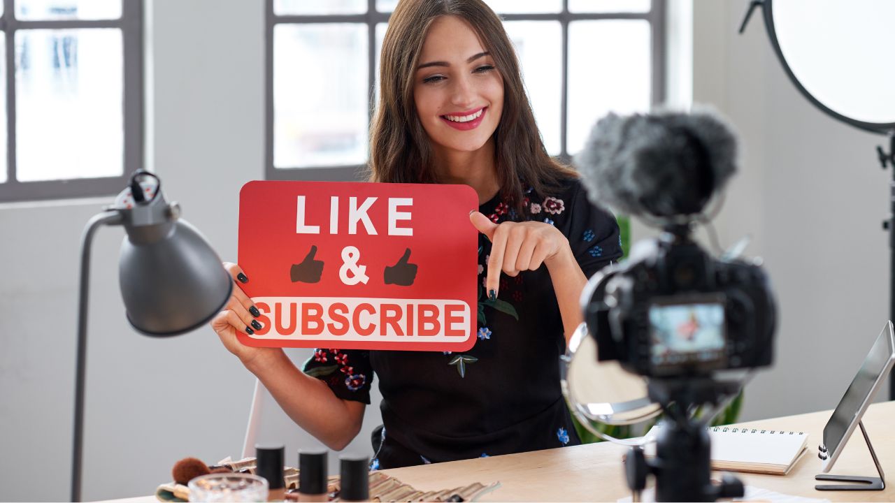 Lächelnde junge Frau mit langen braunen Haaren hält ein rotes Schild mit der Aufschrift ›LIKE & SUBSCRIBE‹ und zwei Daumen-hoch-Symbolen hoch und zeigt mit dem Zeigefinger darauf. Sie sitzt vor einer Kamera mit Mikrofon in einem hellen Studio.