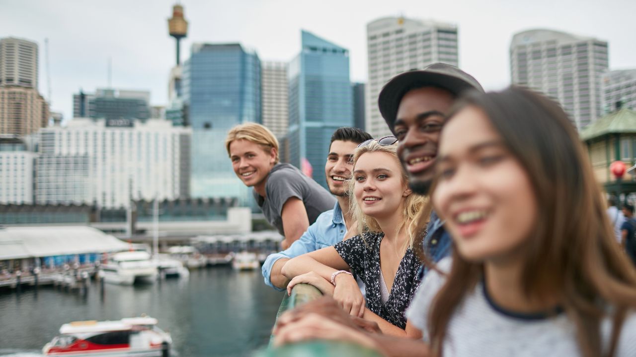 Fünf junge Erwachsene lehnen lächelnd an einer Uferbrüstung und blicken auf einen Hafen mit Booten; im Hintergrund die Skyline von Sydney.