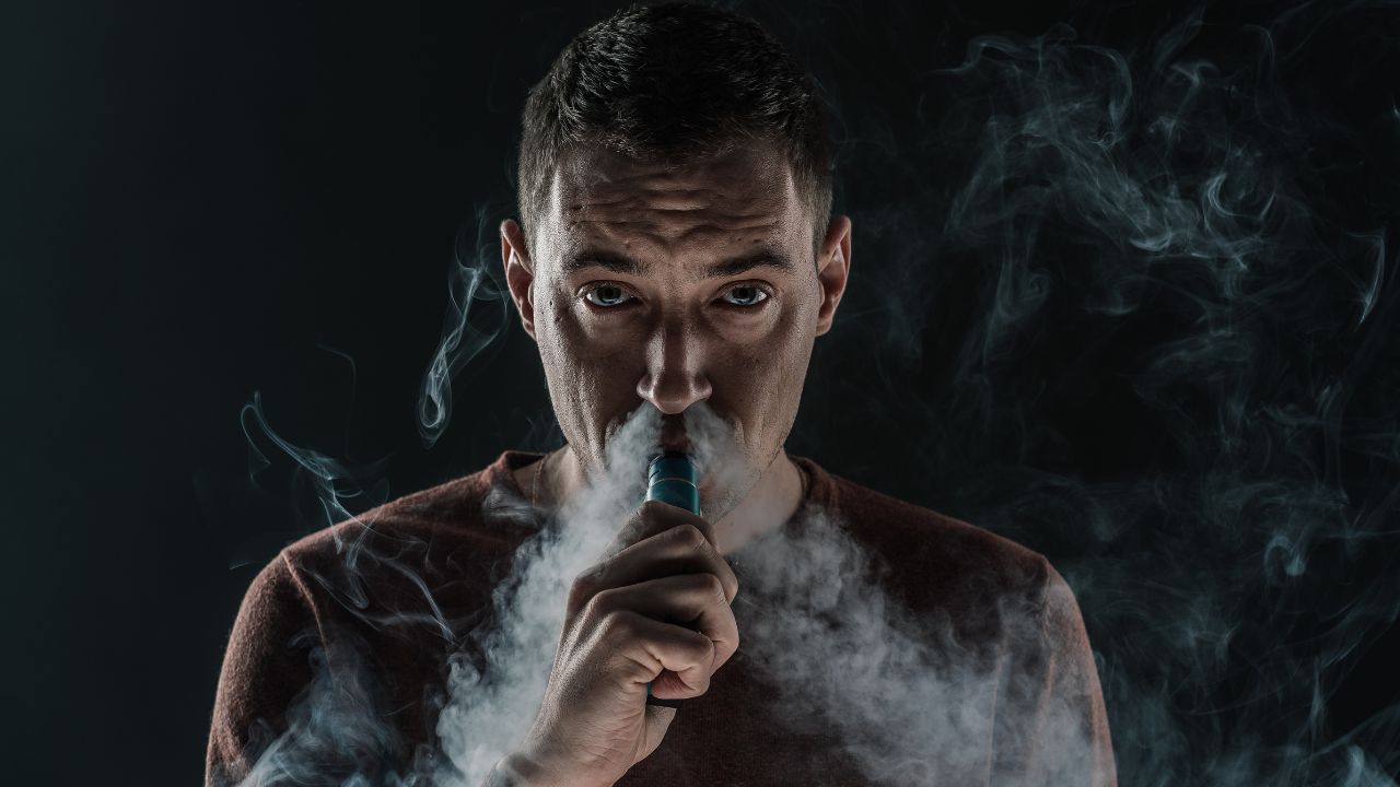 Ein Mann mit kurzen dunklen Haaren und ernstem Gesichtsausdruck hält ein blaues Vape-Gerät an den Mund und atmet eine große, dichte Wolke aus weißem Dampf aus. Er trägt ein rotbraunes T-Shirt vor einem komplett schwarzen Hintergrund, wodurch der Dampf stark kontrastiert.