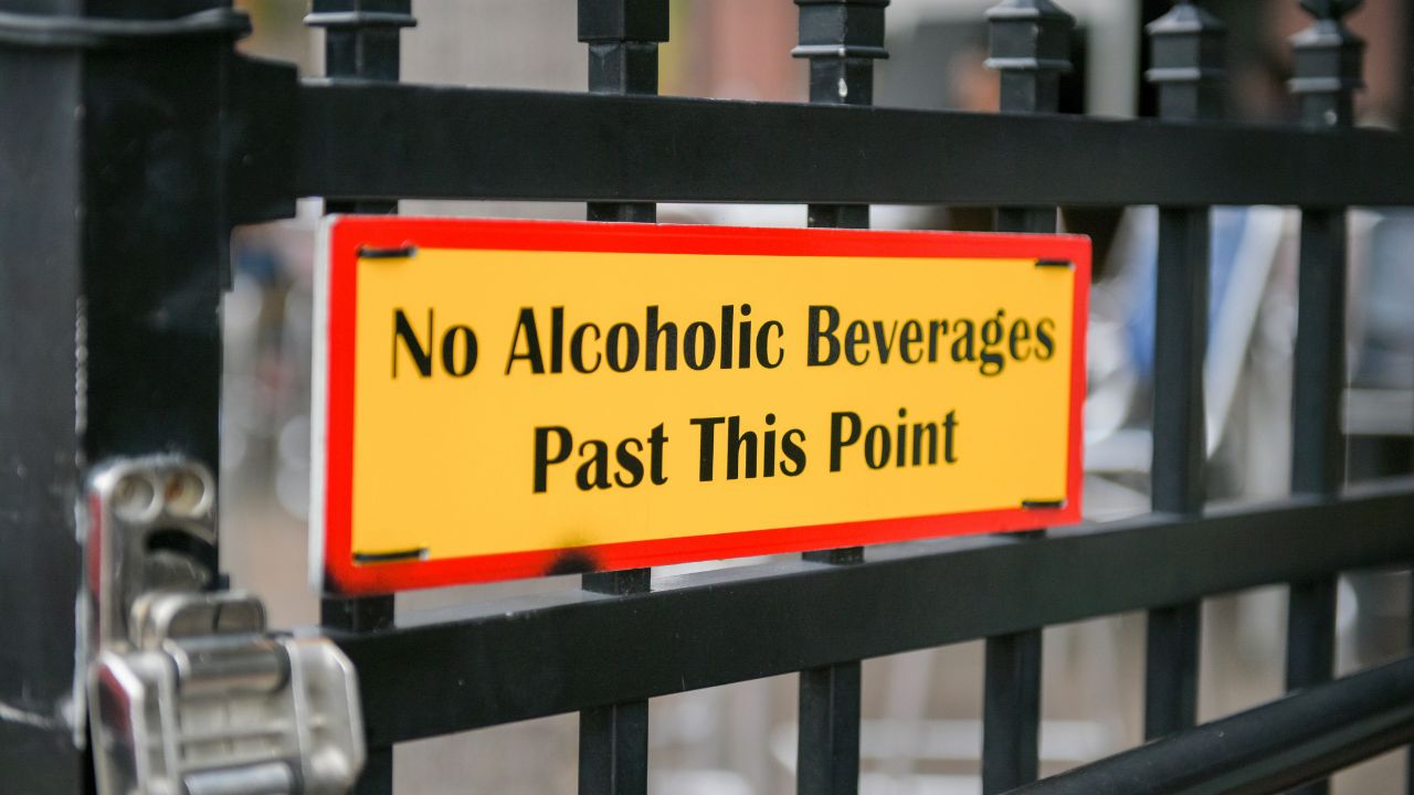 Nahaufnahme eines gelben Schildes mit rotem Rand, das an einem schwarzen Metalltor befestigt ist. Auf dem Schild steht in schwarzen Buchstaben der englische Text: 'No Alcoholic Beverages Past This Point', zu deutsch 'Keine alkoholischen Getränke ab hier'.