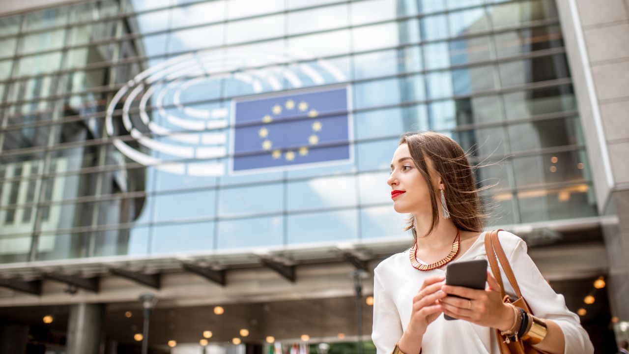 Elegant gekleidete junge Frau in weißer Bluse hält ein Smartphone in der Hand und blickt zur Seite; im Hintergrund die verglaste Fassade des Europäischen Parlaments mit dem blauen EU-Sternenkreis-Emblem.