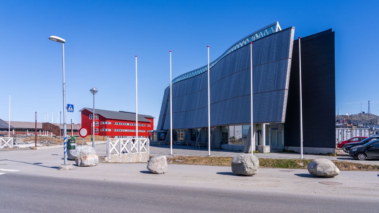 Katuaq, das Kulturzentrum in Grönlands Hauptstadt Nuuk: Modernes, dunkel verkleidetes Gebäude mit geschwungener Dachlinie an einer Straße; davor Fahnenmasten, große Steine und im Hintergrund ein rotes Haus unter klarem blauem Himmel.