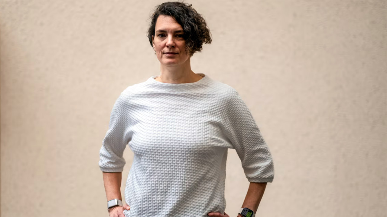 Kristína Šperková, Präsidentin von Movendi Schweden, in einem weißen strukturierten Pullover, steht selbstbewusst mit den Händen auf den Hüften vor einer hellen Wand. Sie trägt eine Smartwatch am linken Handgelenk.